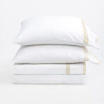 Stack of white pillows stacked on top of each other with beige seam tape against a plain white background conveying a clean bedding scene