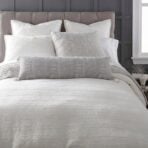 Close up of a neatly made bed with white pillows and a textured gray throw against a dark gray paneled headboard and a window on the right side
