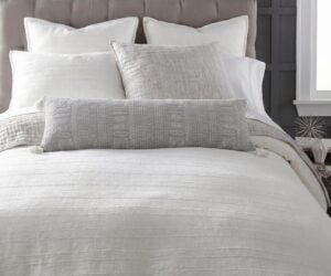 Close up of a neatly made bed with white pillows and a textured gray throw against a dark gray paneled headboard and a window on the right side