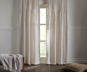 Cream colored curtains frame a tall window in a bright minimalist bedroom a light blanket rests on a wooden bench to the left and a woven floor pillow sits on the dark wood floor to the right