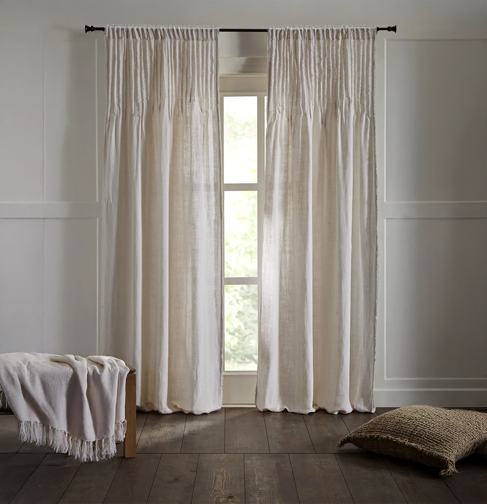 Cm15439w | Amity Home Cream colored curtains frame a tall window in a bright minimalist bedroom a light blanket rests on a wooden bench to the left and a woven floor pillow sits on the dark wood floor to the right
