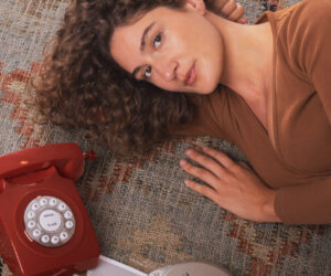 Woman lying on a patterned rug beside a red rotary telephone and magazines looking up at the camera with a calm expression
