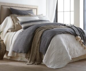 Cozy bedroom scene with a made bed layered gray and white bedding knitted throw and a potted plant on a side table nearby
