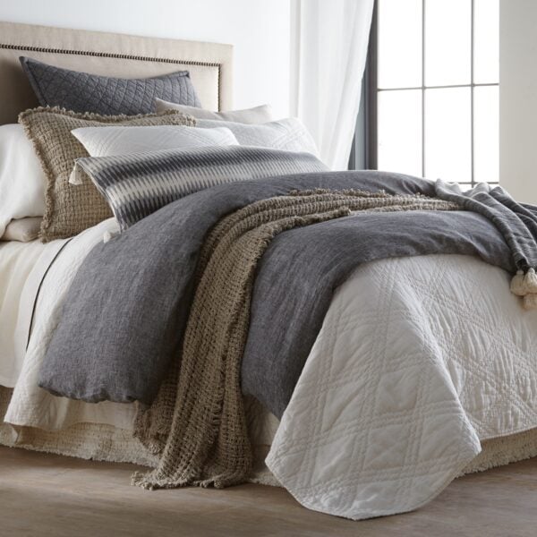 Cozy bedroom scene with a made bed, layered gray and white bedding, knitted throw, and a potted plant on a side table nearby