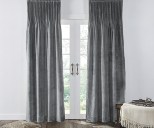 Gray velvet curtains frame white french doors with a view of greenery outside a woven floor pillow sits nearby and a fringed throw rests on a bench to the right