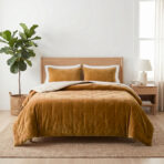 Wooden bed with mustard velvet bedding and two matching pillows a tall green plant in a woven basket sits to the left a nightstand and lamp on the right in a bright minimalist room