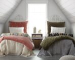 Twin beds with white ornate metal headboards coral and olive throw pillows in a bright attic bedroom by a window between the beds