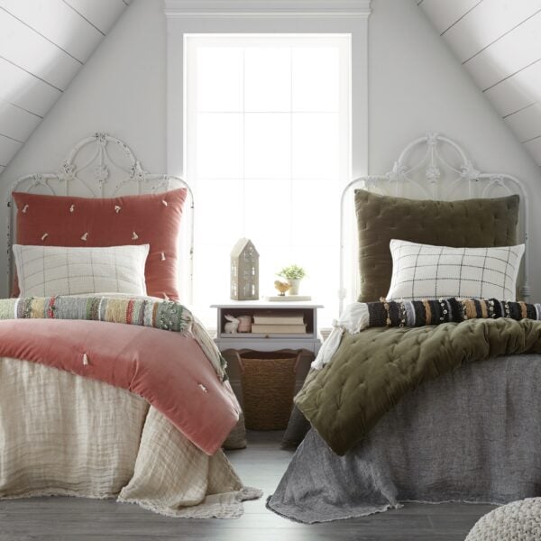 Twin beds with white ornate metal headboards, coral and olive throw pillows in a bright attic bedroom by a window between the beds.