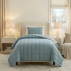 Twin bed with a blue quilted bedspread and matching pillow flanked by white nightstands and lamps in a bright bedroom
