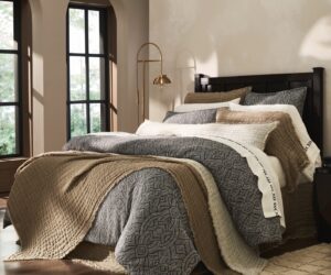 A cozy bedroom with a dark wood bed beige and patterned gray bedding and a brass arc floor lamp beside the bed near large arched windows with sheer curtains
