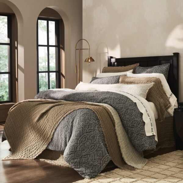A cozy bedroom with a dark wood bed, beige and patterned gray bedding, and a brass arc floor lamp beside the bed, near large arched windows with sheer curtains.