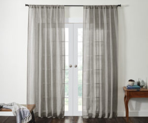 Grey sheer curtains frame white french doors with a view of greenery outside A wooden bench with a throw sits to the left and a small table with books and decor to the right