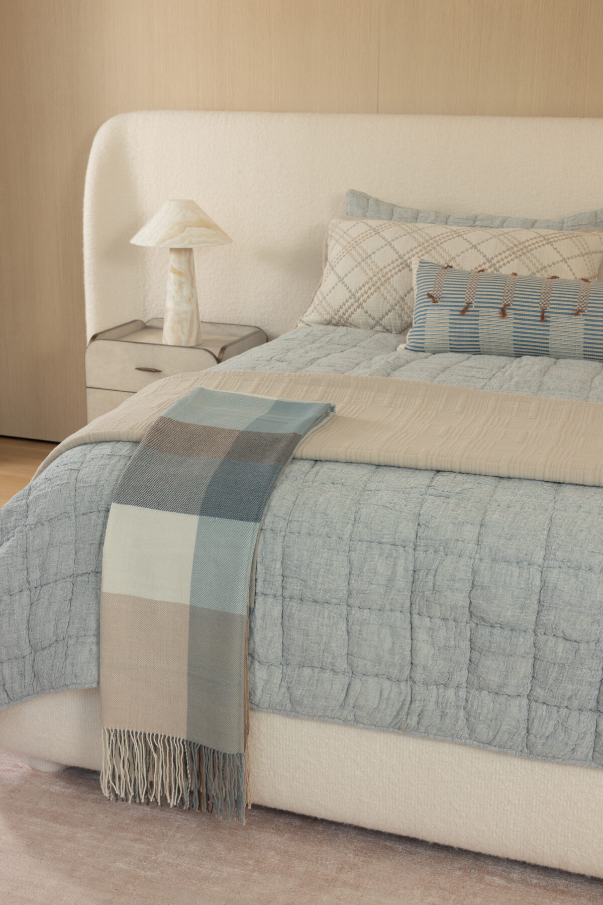 Cozy bedroom scene with a tufted headboard blue quilt striped and plaid pillows and a marble bedside lamp on a nightstand fringed throw hangs over the edge of the bed