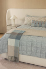 Cozy bedroom scene with a tufted headboard blue quilt striped and plaid pillows and a marble bedside lamp on a nightstand fringed throw hangs over the edge of the bed