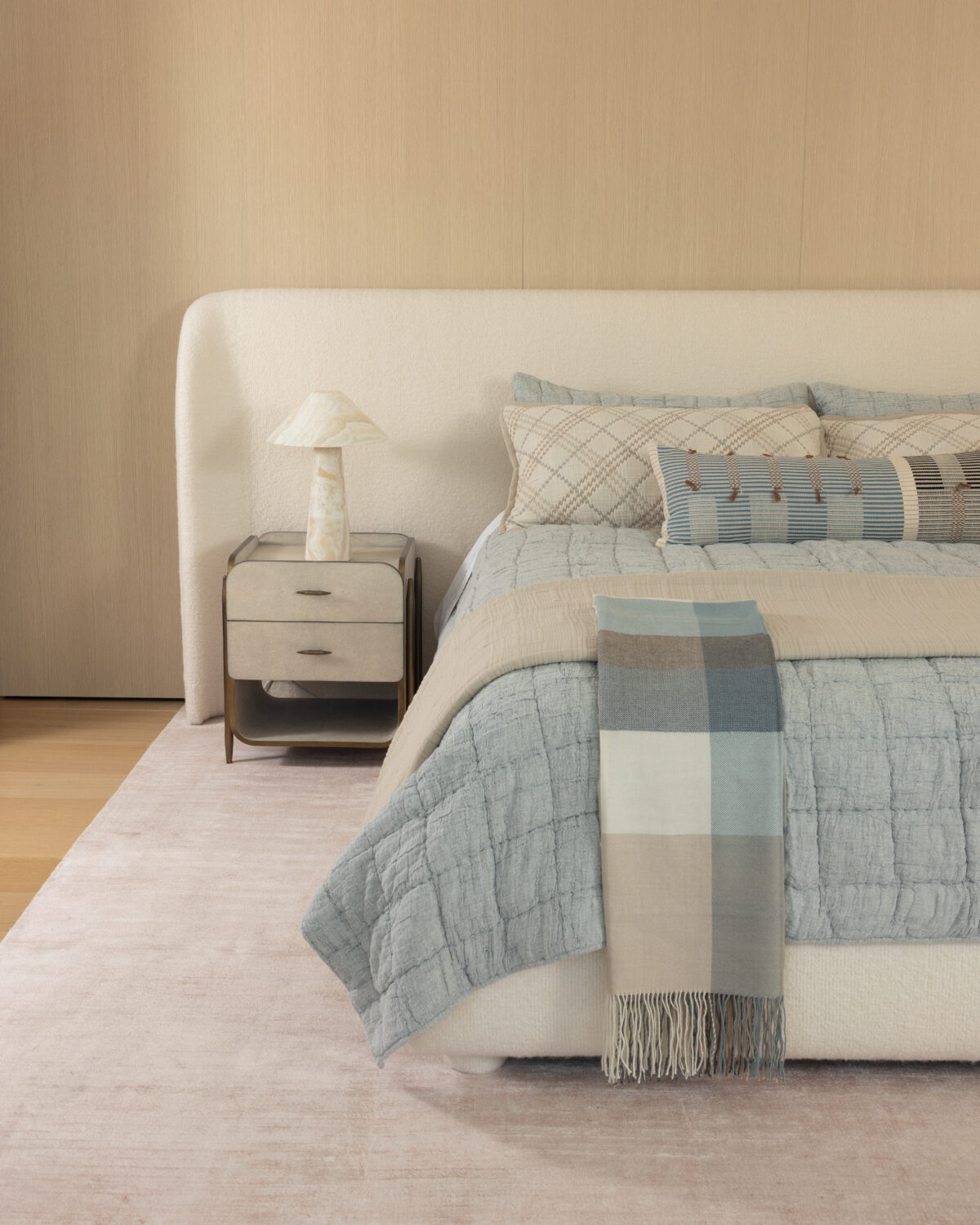 Neutral toned bedroom with a white upholstered headboard nightstand and a blue gray quilted bed with plaid pillows and a fringed throw scarf