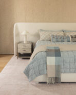 Neutral toned bedroom with a white upholstered headboard nightstand and a blue gray quilted bed with plaid pillows and a fringed throw scarf