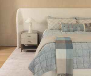 Neutral toned bedroom with a white upholstered headboard nightstand and a blue gray quilted bed with plaid pillows and a fringed throw scarf