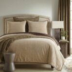Beige upholstered bed with studded trim matching pillows and a textured throw a round gray stool sits at the foot of the bed bedside table with a lamp is visible on the right