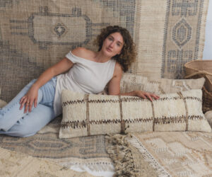 Young woman with curly hair lying down on a layered beige and blue woven rug setup modeling casually