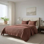 Bedroom scene with a bed covered in dusty rose bedding white headboard and a white curtained window letting in natural light potted plant on the sill nearby