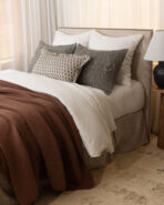 Bed with beige upholstered headboard and multiple pillows brown throw blanket in a softly lit bedroom