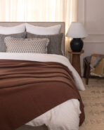Bed in a cozy bedroom with white sheets brown blanket and decorative pillows by a tufted headboard A lamp sits on a wooden nightstand to the right