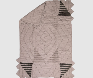 Soft pink quilt with a diamond quilted pattern jagged fringe edges along the borders and black stepped chevron motifs in the corners