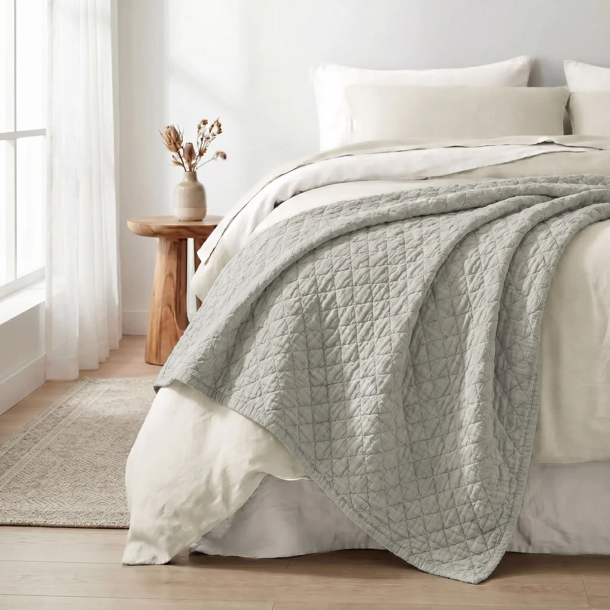 Neatly made bed with a light gray quilt white sheets and pillows beside a wooden nightstand with a beige vase of dried flowers