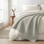 Neatly made bed with a light gray quilt white sheets and pillows beside a wooden nightstand with a beige vase of dried flowers