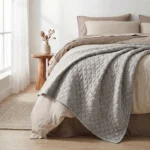 A neatly made bed with quilted gray bedding and beige pillows beside a wooden nightstand with a vase in a bright bedroom