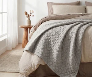 A neatly made bed with quilted gray bedding and beige pillows beside a wooden nightstand with a vase in a bright bedroom