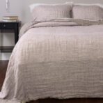 Bed with beige linen sheets and pillows a black nightstand with books beside it and a hanging bulb light
