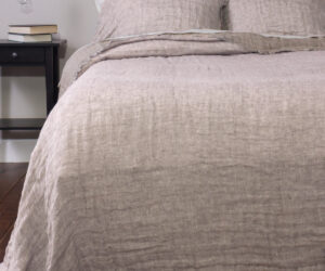 Bed with beige linen sheets and pillows a black nightstand with books beside it and a hanging bulb light