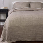 Bed with beige linen bedding and pillows a dark nightstand with books beside it and a hanging light bulb above the table