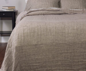 Bed with beige linen bedding and pillows a dark nightstand with books beside it and a hanging light bulb above the table