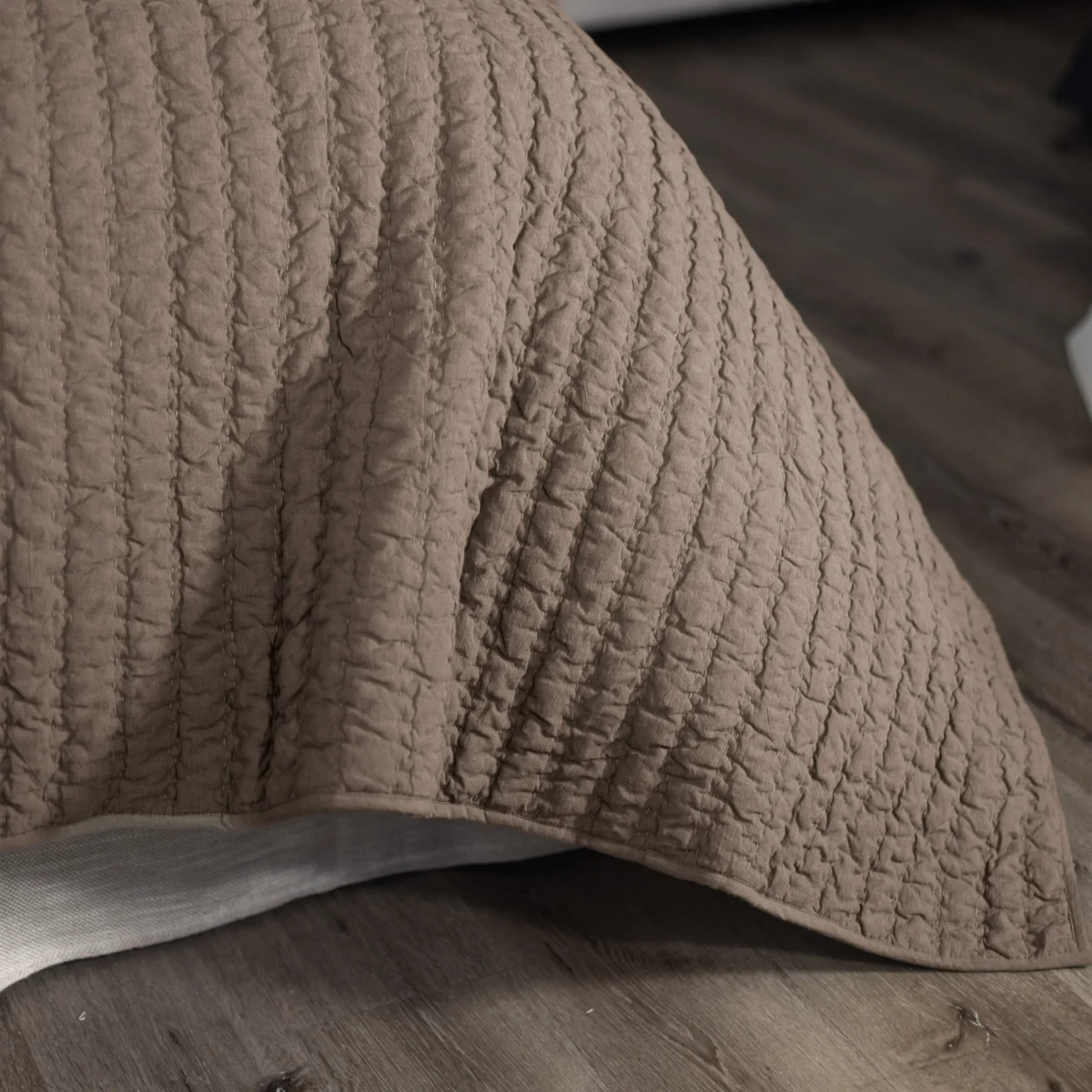 Close up of a beige quilt with vertical stitched channels draped over a bed showing textured fabric