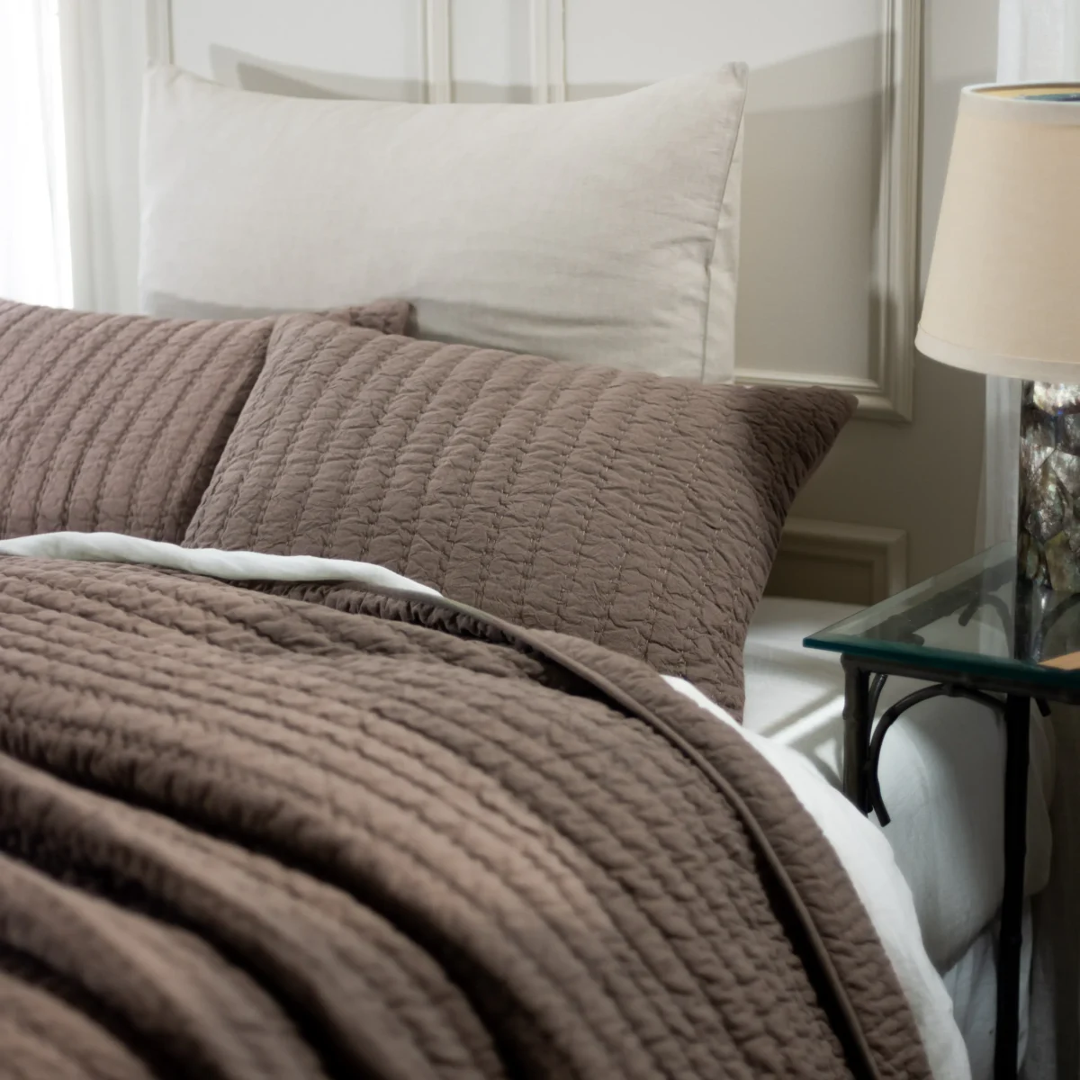 Bed made with a brown quilted coverlet and white pillows in a cozy bedroom with a glass side table and lamp nearby