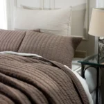 Bed made with a brown quilted coverlet and white pillows in a cozy bedroom with a glass side table and lamp nearby