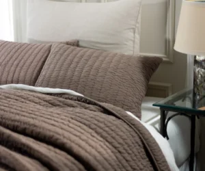 Bed made with a brown quilted coverlet and white pillows in a cozy bedroom with a glass side table and lamp nearby