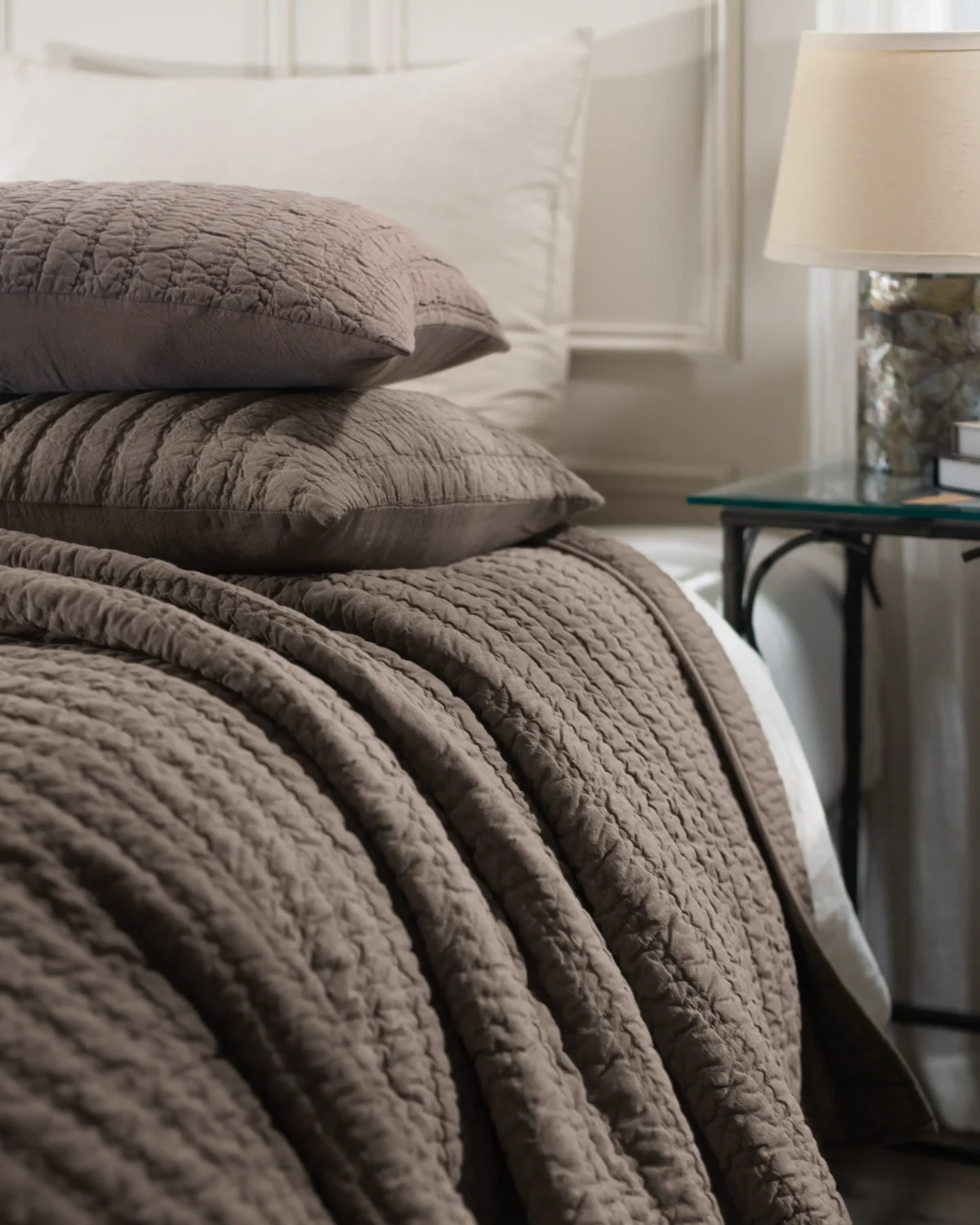 Brown quilted bedding stacked on a bed with a white headboard and glass topped side table in the background