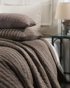 Brown quilted bedding stacked on a bed with a white headboard and glass topped side table in the background