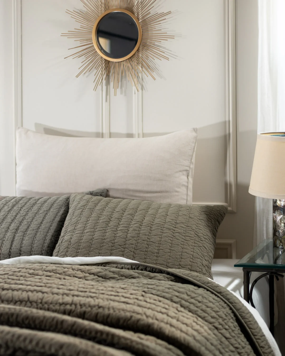 Gray quilted bedspread with a white pillow and a gold sunburst mirror on the wall above the headboard creating a luxe bedroom detail