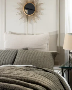 Gray quilted bedspread with a white pillow and a gold sunburst mirror on the wall above the headboard creating a luxe bedroom detail