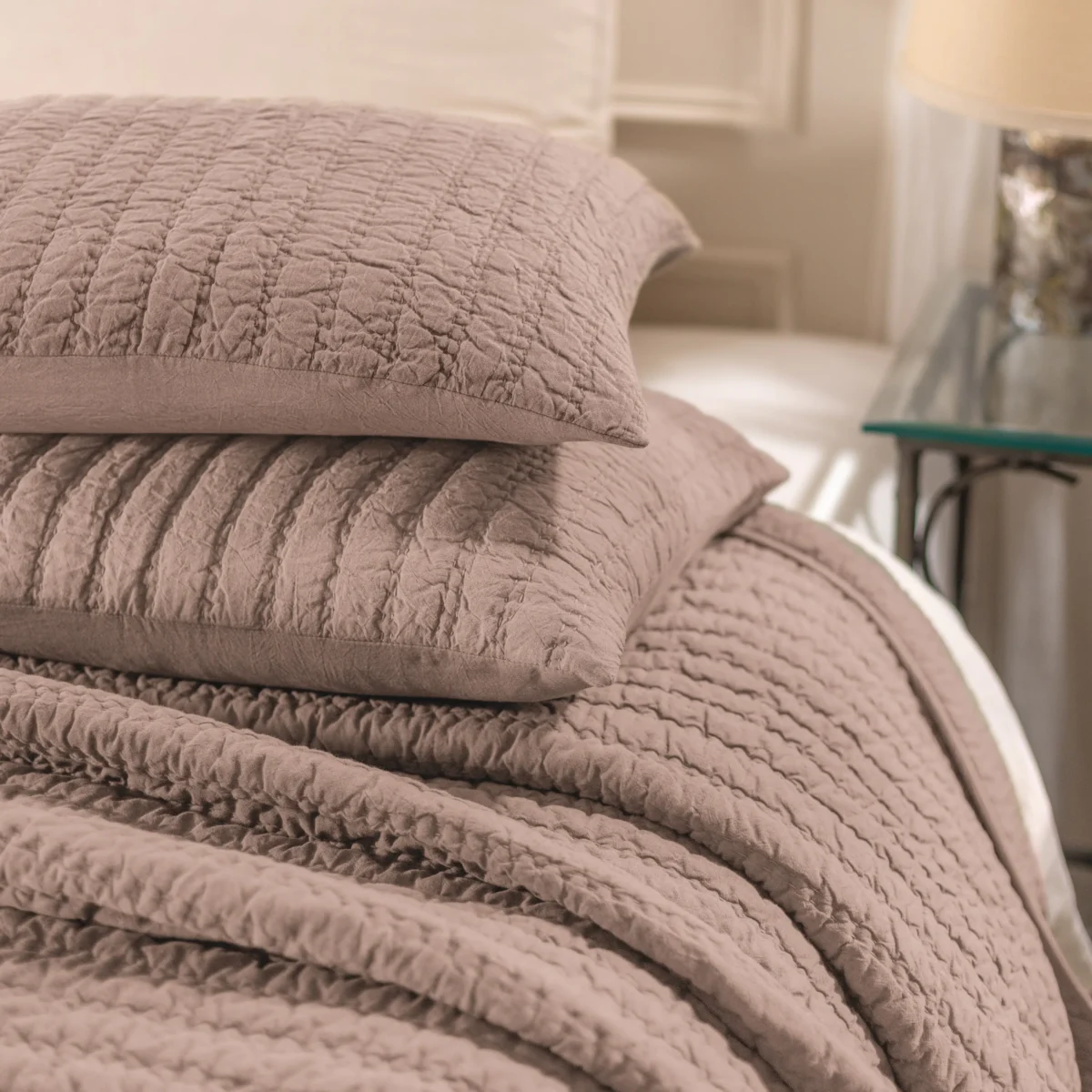 Close up of stacked quilted blankets in dusty rose on a bed showing textured stitching and soft fabric folds
