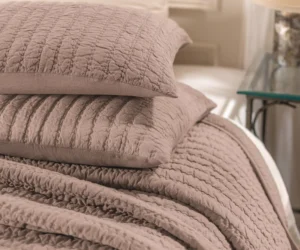 Close up of stacked quilted blankets in dusty rose on a bed showing textured stitching and soft fabric folds