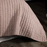 Close up of a soft pink quilt with vertical stitched channels laid on a dark wooden surface prop or table edge visible at the bottom right