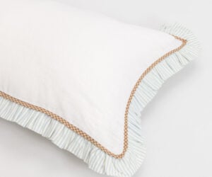 Close up of a white pillow with a blue striped pillow underneath and brown trim along the edge showing overlapping fabrics