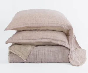 Stack of several light beige linen pillows with frayed edges arranged against a white backdrop showcasing soft textured fabric