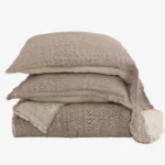 Stack of beige linen pillows with frayed edges arranged in a tower against a white background textured fabric visible