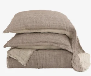 Stack of beige linen pillows with frayed edges arranged in a tower against a white background textured fabric visible